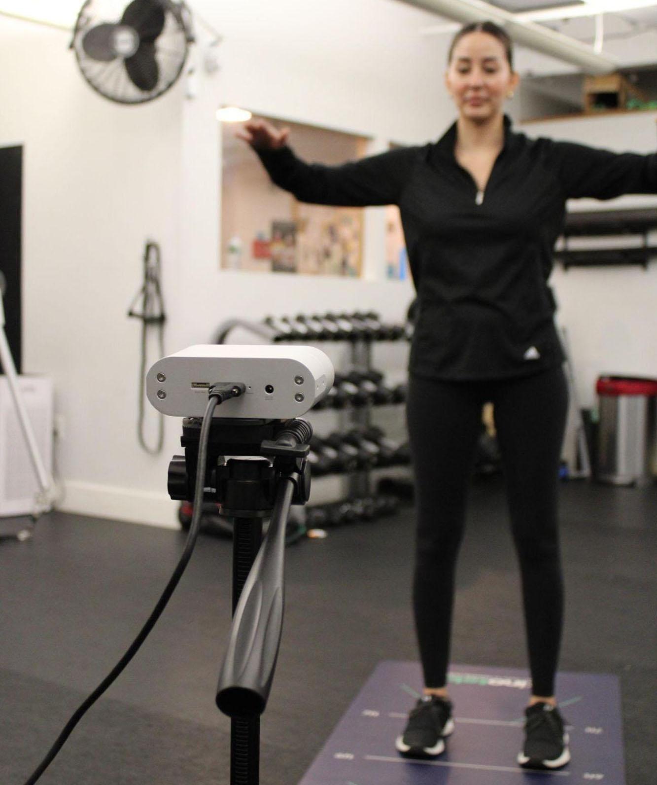 a woman in athletic wear stands on a mat in a gym arms outstretched in front of a camera on a tripod